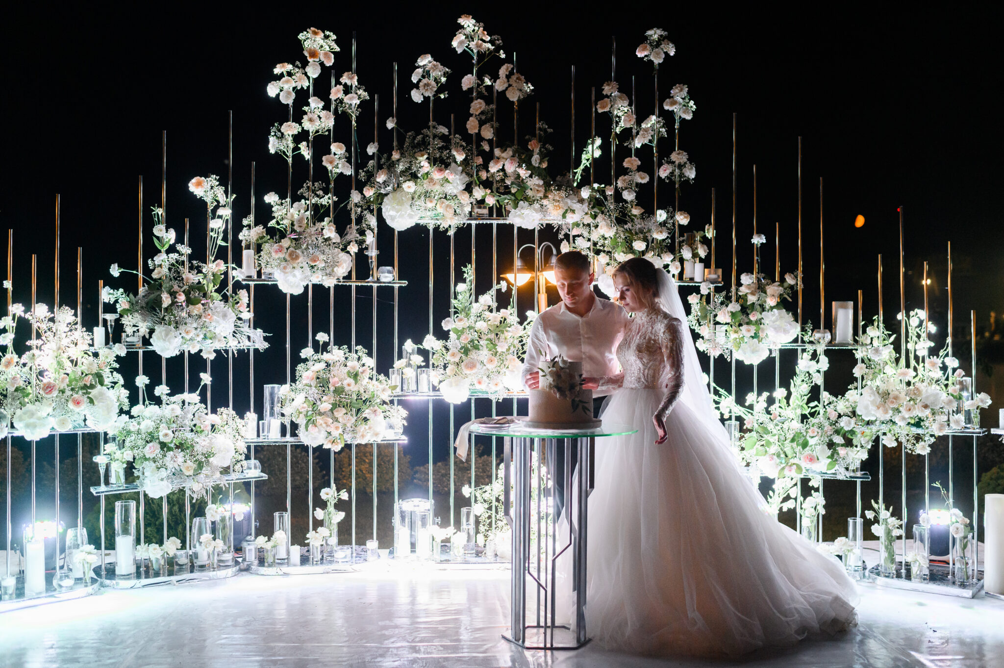 Side view of loving brides couple cutting flowering white cake on evening wedding ceremony, woman wearing in beautiful puffy dress and man in white shirt,standing near lighting floral arch,