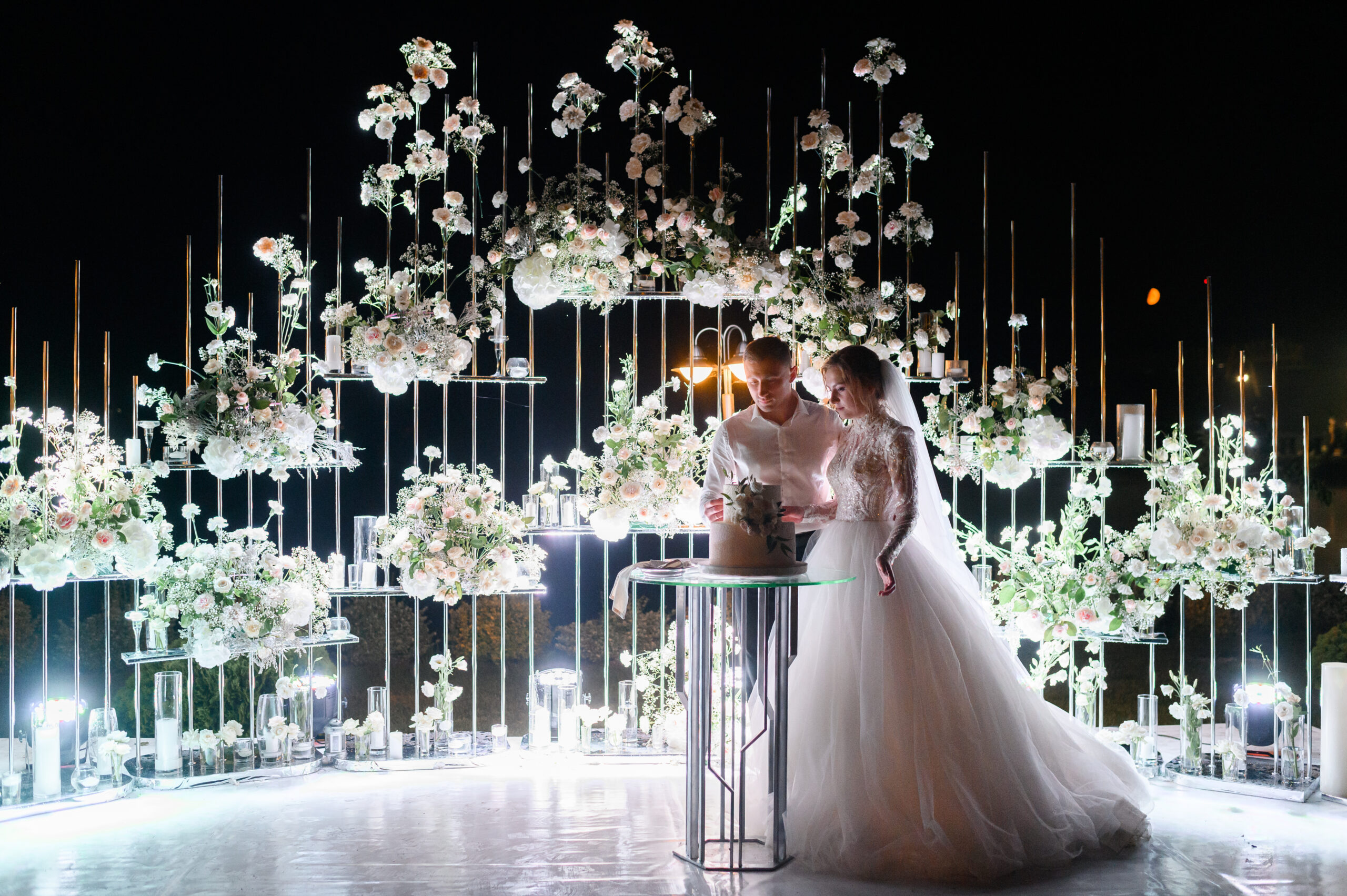 Side view of loving brides couple cutting flowering white cake on evening wedding ceremony, woman wearing in beautiful puffy dress and man in white shirt,standing near lighting floral arch,